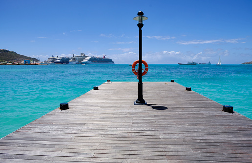 Saint Maarten — March 28, 2019: Pier extends into the turquoise water of the Caribbean. A life preserver is hanging on a light pole. Cruise ships are at a dock in the distance.