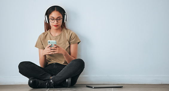 Music headphones, floor and woman with phone in home by wall background with mockup. Cellphone, social media and female sitting on ground while streaming or listening to podcast, radio sound or audio