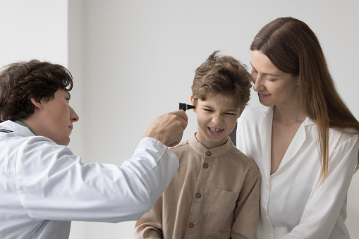 Focused doctor checking ears of naughty boy for infection, using otoscope tool. Patient kid and mom visiting family doctor office for regular healthcare medical examination