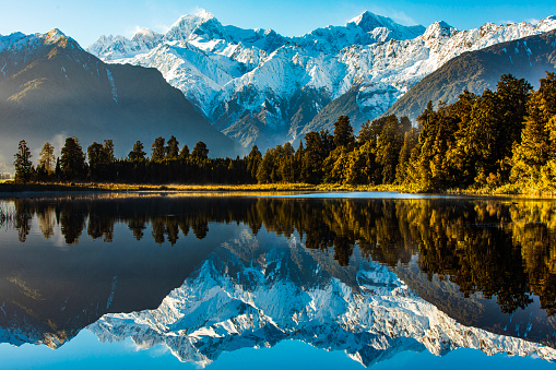 Reflection of snow capped mountains on a still lake with sun shining on forest, shot in New Zealand.