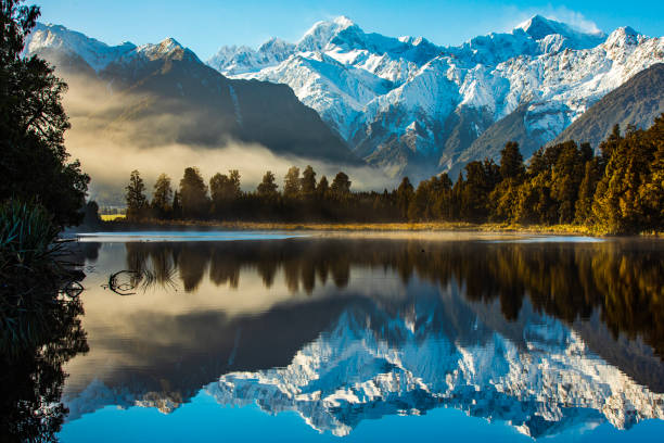 Reflection of snow capped mountains on a still lake with sun shining on forest Reflection of snow capped mountains on a still lake with sun shining on forest, shot in New Zealand. winter sunrise mountain snow stock pictures, royalty-free photos & images