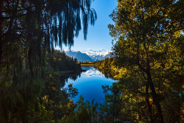 istock Reflection of snow capped mountains on a still lake with sun shining on forest framed by trees 1497730925