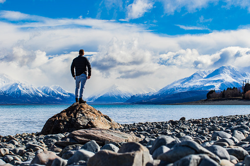 Male model standing in sunlight on lakeshore with snow covered mountains in background, lifestyle shoot in New Zealand.