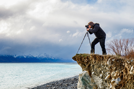 Young professional man operating camera equipment on tripod setup taking photographs of dramatic mountain landscape in New Zealand in golden light.