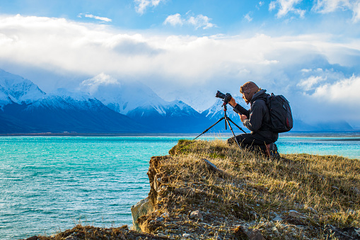 Young professional man operating camera equipment on tripod setup taking photographs of dramatic mountain landscape in New Zealand in golden light.