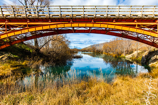 Old bridge over turquoise blue green river with orange foliage in the mountains of New Zealand.