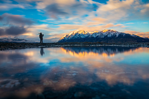 Young male professional photographer contemplating the view of snow capped mountain range on a still lake at sunset in New Zealand.