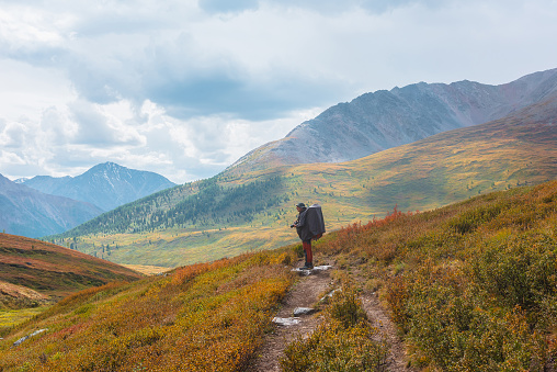 Alone traveler in raincoat with large backpack photographs nature among fading autumn vegetations on hiking trail through mountain pass. Backpacker with photo camera shoots autumn mountain landscape.