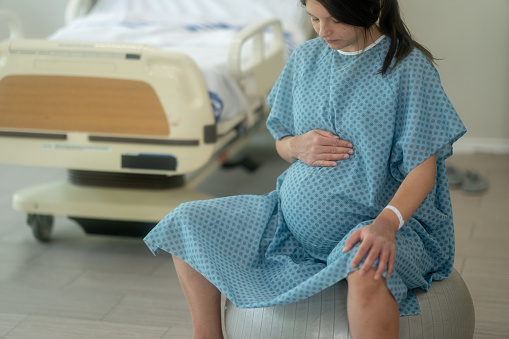 A pregnant woman sits on a birthing ball as she tries to relieve the pain by bouncing through her contractions and alleviating pressure in her pelvis.  She is wearing a blue hospital gown and is focused on her breathing.