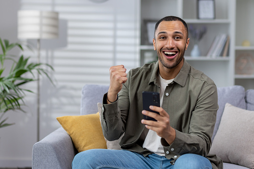 Portrait of a happy young African American man sitting on the couch at home and looking at the camera while using the phone. Celebrating victory, winning.