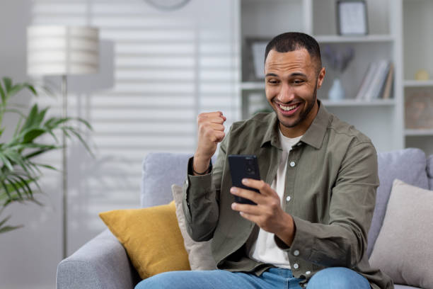 istock Young smiling Muslim man using phone while sitting at home on sofa, playing online games, placing bets, enjoying money win, celebrating, showing victory gesture with hand 1497754056