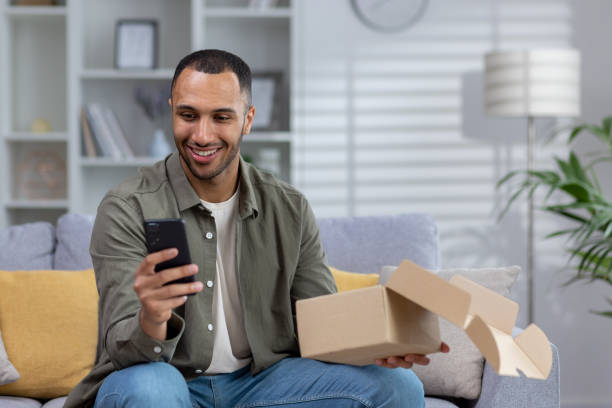 istock Satisfied Latin American man sits at home on the sofa and holds, opens the package he received home delivery by courier. Uses the phone, makes an online order through the application 1497754573
