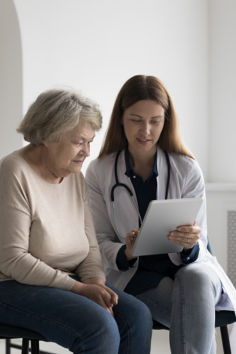 Friendly positive GP doctor showing tablet screen to elderly 80s patient woman, explaining examination result, treatment, roentgen electronic shot, giving consultation, expertise on appointment