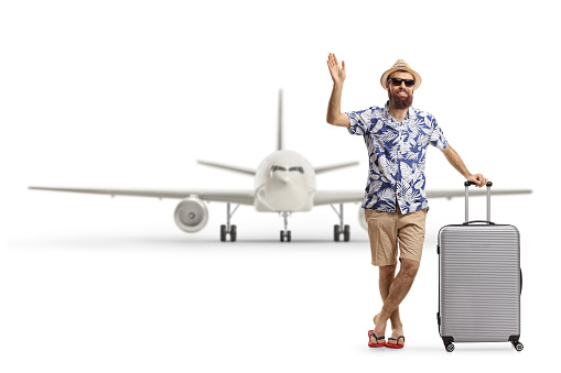 Full length portrait of a bearded man tourist posing with a suitcase and waving in front of an airplane isolated on white background
