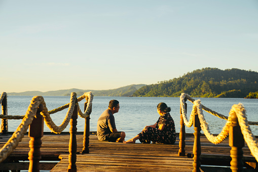 asian couple sitting at the edge of a jetty at sunset in terusan mande, painan, west sumatra