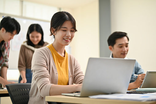 Close-up image of a beautiful and smart young Asian female worker in a startup team is working on her tasks on hera laptop while her colleagues are in the room.