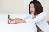 closeup beautiful asian woman, short hair in white shirt using video call or self portrait from mobile phone on white bed in bedroom with happy smiling face