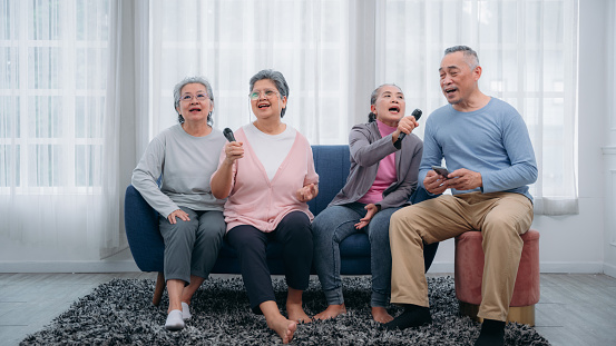 Senior citizens performing karaoke. Senior Asian friends singing karaoke on a sofa in the living space with a smile and a joyful look. Buddies enjoying karaoke in their homes.