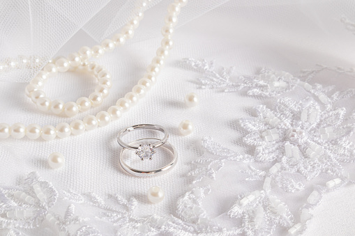 Gentle wedding background. Two white gold engagement rings with a diamond on a white embroidered veil and a pearl necklace. space for text