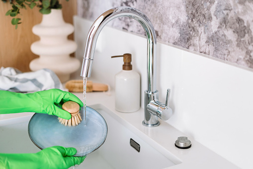 cropped shot of female hand in rubber gloves wash dish with wooden brush at sink with stainless steel faucet at modern kitchen, zero waste concept