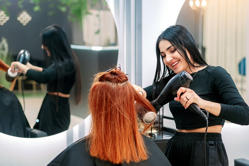 Hair styling in beauty salon. Woman does her hair in modern beauty salon. Woman stylist dries hair with hairdryer to salon client and smiles.