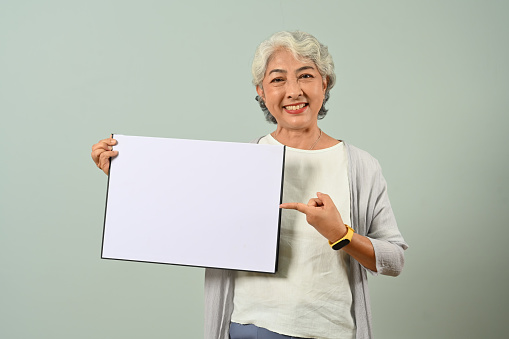 Portrait of smiling gray-haired 60s woman holding blank advertising board standing over light blue background.