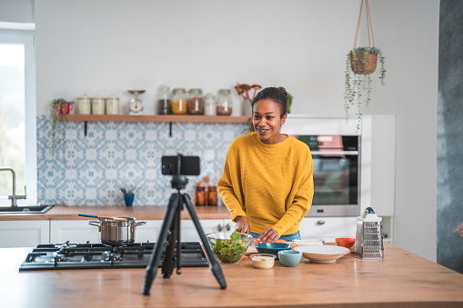 A joyful food vlogger filming herself in her home kitchen using a mobile phone camera. She guides her social media followers through the process of preparing a wholesome meal rich in vegetables.