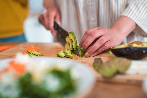 Close-up photo of an unrecognizable woman expertly slicing ingredients with a kitchen knife for a wholesome vegetable meal. She precisely cuts avocado into slices and prepares other components for vibrant salad.