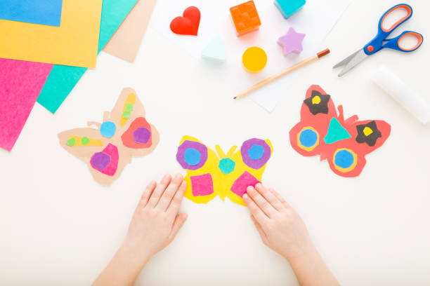 istock Little child hands holding colorful butterfly shape from paper on white table background. Making decoration elements. Closeup. Point of view shot. Top down view. 1497817846
