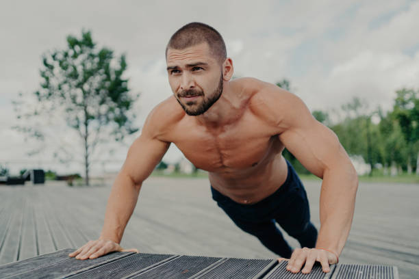 l'uomo barbuto a torso nudo fa flessioni nel parco, allena la parte superiore del corpo, mantiene una routine di allenamento regolare. atleta motivato in azione. - macho foto e immagini stock