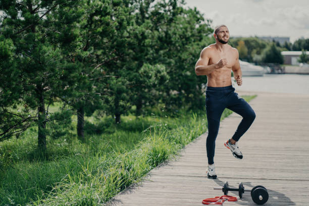 Active man jumps outdoors, warms up for jogging, wears sport trousers and sneakers, exercises with bare torso. Active man jumps outdoors, warms up for jogging, wears sport trousers and sneakers, exercises with bare torso. Barbell stock pictures, royalty-free photos & images