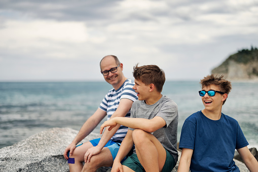 Father and two teenage boys enjoying spring day in Calabrian town of Tropea. They are sitting on the rocks on beach, talking and laughing. \nSpringtime, off-season vacations day in Tropea, Catania, Italy.\nShot with Canon R5