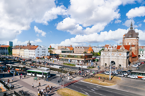 This large panorama photograph captures the vibrant cityscape of Gdansk, Poland, on a sunny day with a picturesque blue sky adorned with fluffy clouds. Taken from the viewpoint of the Forum shopping mall, the view encompasses the old city, with prominent landmarks such as Wyżynna Gate (Brama Wyżynna) and Długouliczna Gate (Brama Długouliczna) showcased in the frame. The street below is bustling with cars and various forms of public transport, while pedestrians traverse the vibrant pedestrian areas. With its non-fictional depiction, this photograph presents an authentic portrayal of Gdansk's urban environment, offering a captivating scene accentuated by the delightful blue sky and fluffy clouds.