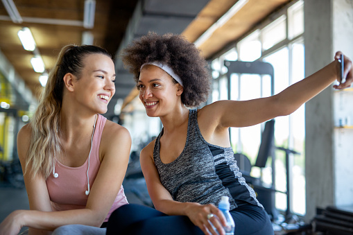 Happy fit women, friends smiling, talking and taking photos after work out in gym. Social media, people, sport concept.
