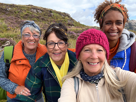 A mobile shot of a group of senior female Friends taking a selfie together smiling into the camera. They are standing near the village of Diabaig in the west highlands of Scotland with a mountain range behind them.