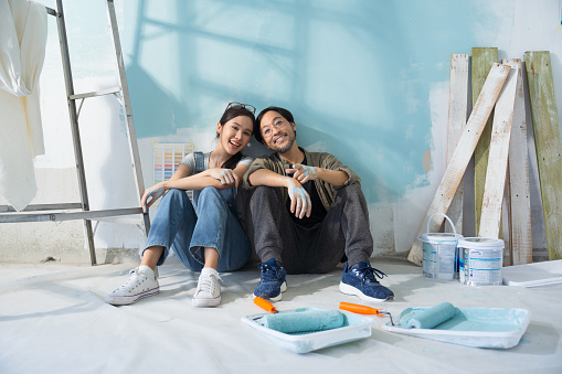 Portrait Asian couple repairing and painting the wall with blue paint using a roller during renovation in their new apartment.