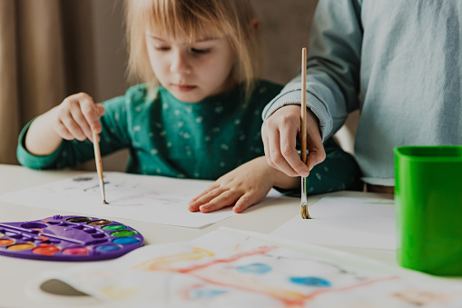 Two cute little sisters, classmates are painting on table. Small girl kids with brush in school. Happy adorable preschooler, 4 and 7 years child in living room. Home schooling concept, doing homework