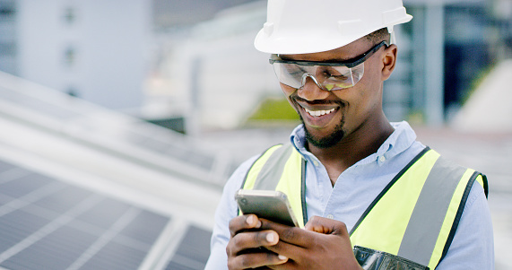 A young man using a smartphone while installing solar panels on the roof of a building, A construction worker working online with his phone on a renewable energy project