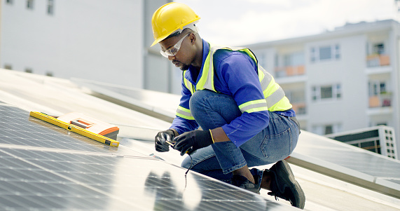 A young man using a drill, measuring tape and spirit level while installing solar panels on the roof of a building. A man working with tools on a renewable energy project