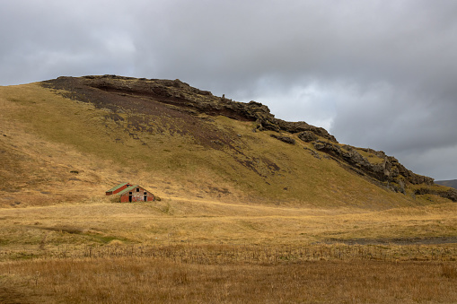 Small traditional original house on a pasture, under a mountain. Yellow color of the grass in the autumn. Cloudy sky. South-central Iceland.