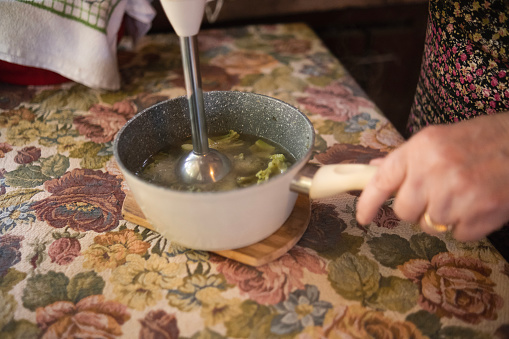 Happy grandmother using a kitchen utensil to blend broccoli into a delicious creamy soup