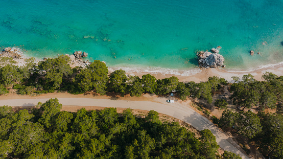 Drone photo of the car camping by the beautiful road at the peninsula surrounded by the green tree area and the crystal blue Mediterranean Sea at the coastal Turkey
