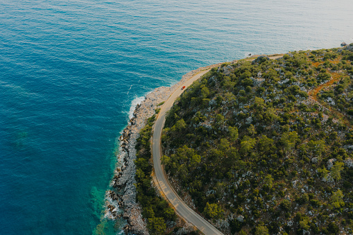 Drone photo of the car driving the beautiful road at the peninsula surrounded by the green tree area and the crystal blue Mediterranean Sea at the coastal Turkey
