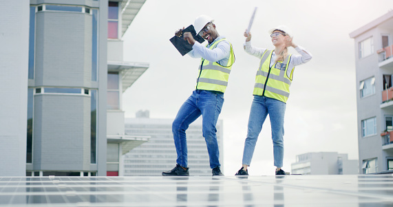 A young man and woman dancing while installing solar panels on the roof of a building. Diverse team having fun while working on a renewable energy building project