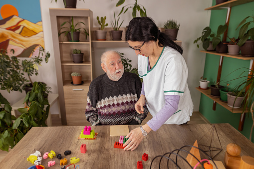 A female nurse is helping a senior man with dementia play with colorful toys during a toy therapy session.