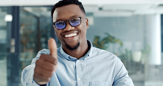 Happy and confident young businessman showing a thumbs up in a modern office. Portrait of a man making a hand sign of to show support. Male businessperson smiling and looking pleased