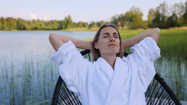 woman in white bathrobe relaxing in chair with eyes closed on lake footbridge at warm summer evening. mental health and wellness