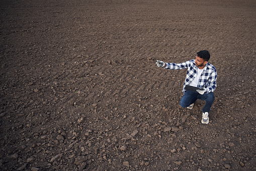 Cultivated agricultural field. Sitting, holding digital tablet. Handsome Indian man.