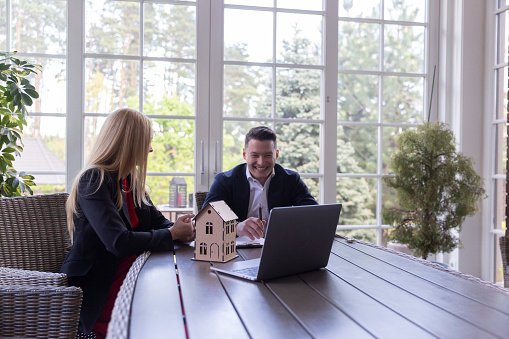 Client and real estate agent smiling during conversation about choosing residence. Professional estate agent shows available variants on laptop