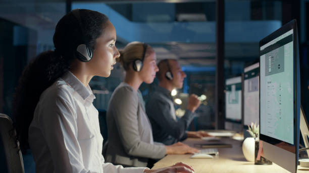 istock Call centre agents talking on headsets working on computers in an office. Female telemarketing consultant operating helpdesk for customer service and sales support during a night shift in an office 1497867895